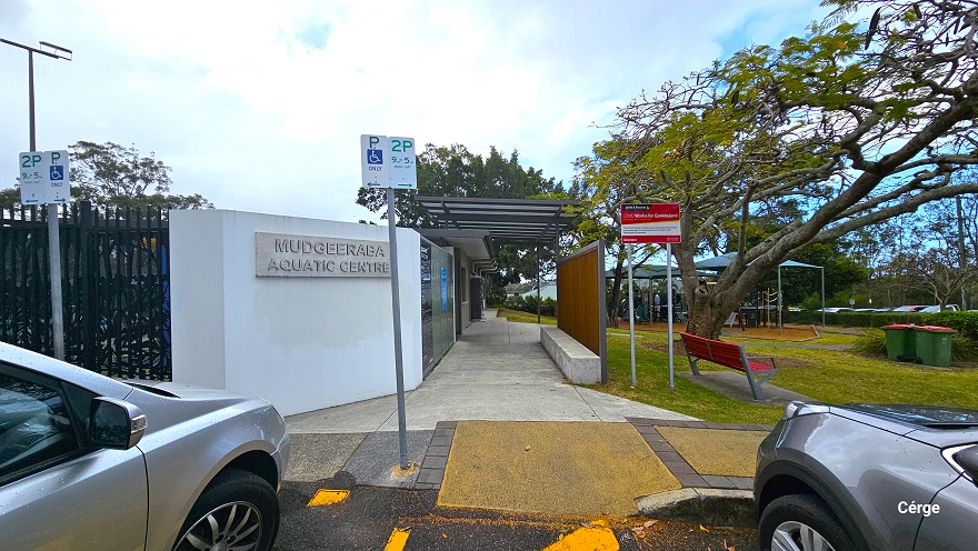 Parking for people with disability is marked by a white parking sign with the letter P, accessibility logo, and the word Only printed on the flat sign board. Beside is the 2-hour parking sign with green print including date details. On the left side is the name of the facility, detailed on a white wall, while on the right is a brown fence with skylit roofing that covers the cemented smooth pathway at the center. At the back of the red bench is a tall red signboard. Parking for people with disability is marked by a white parking sign with the letter P, accessibility logo, and the word Only printed on the flat sign board. Beside is the 2-hour parking sign with green print including date details. On the left side is the name of the facility, detailed on a white wall, while on the right is a brown fence with skylit roofing that covers the cemented smooth pathway at the center. At the back of the red bench is a tall red signboard.