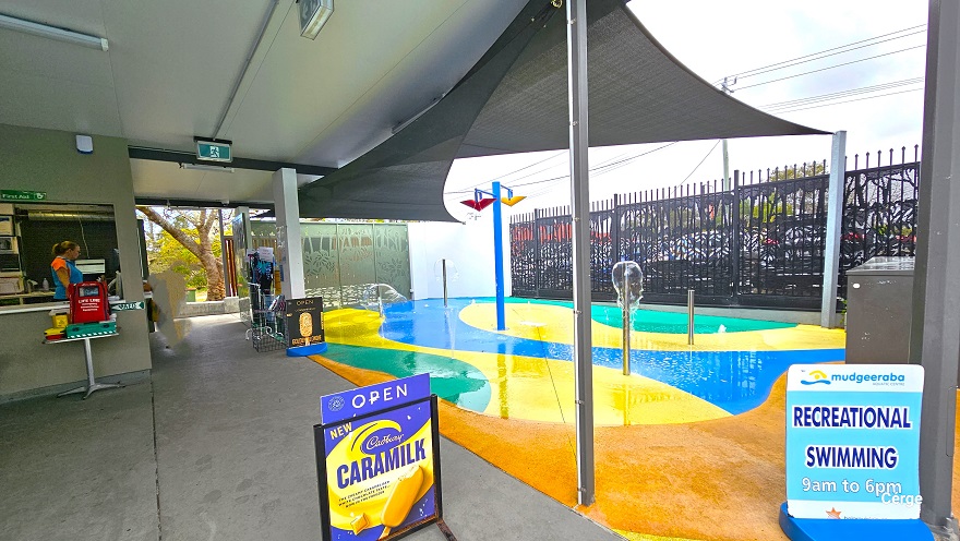 Small children’s splash zone at Mudgeeraba Aquatic Centre can be distinguished by an orange, yellow, blue, and green painted floor directly to the left of the reception. Short metal poles eject water. In the middle of the splash zone is a blue pole with two triangular buckets at the top. Two black sun shades party covers this zone. There are two signboards outside the children’s splash zone. Small children’s splash zone at Mudgeeraba Aquatic Centre can be distinguished by an orange, yellow, blue, and green painted floor directly to the left of the reception. Short metal poles eject water. In the middle of the splash zone is a blue pole with two triangular buckets at the top. Two black sun shades party covers this zone. There are two signboards outside the children’s splash zone.