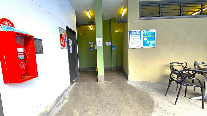 Hallway leading to the bathroom and an accessible bathroom in Mudgeeraba Aquatic Centre. The bathrooms can be distinguished by the lime green painted walls. Mounted on the left white wall is a red box containing the red fire extinguisher. This wall also has a black door. To the right is a seating area with black chairs and a table. The bathrooms are illuminated by a warm fluorescent light. Hallway leading to the bathroom and an accessible bathroom in Mudgeeraba Aquatic Centre. The bathrooms can be distinguished by the lime green painted walls. Mounted on the left white wall is a red box containing the red fire extinguisher. This wall also has a black door. To the right is a seating area with black chairs and a table. The bathrooms are illuminated by a warm fluorescent light.