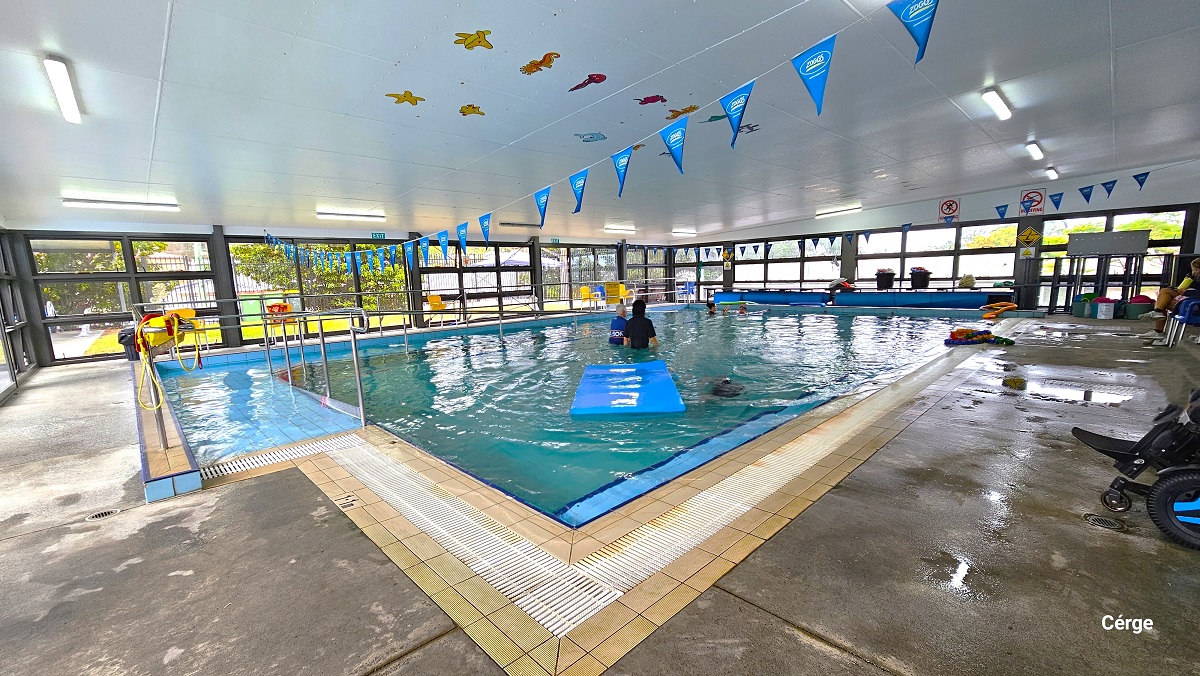 Right next to the tinted glass sliding doors with black frames is the Indoor program pool heated to 33 degrees Celsius and is restricted to rehabilitation and gentle exercise users over the age of 16 in Mudgeeraba Aquatic Centre. The Parent room inside has a wooden bench, a blue plastic picnic table, and a table with a colorful coral structural design. To the back is a glass window and a black door. The Parent room has a smooth cemented floor with a white ceiling. Right next to the tinted glass sliding doors with black frames is the Indoor program pool heated to 33 degrees Celsius and is restricted to rehabilitation and gentle exercise users over the age of 16 in Mudgeeraba Aquatic Centre. The Parent room inside has a wooden bench, a blue plastic picnic table, and a table with a colorful coral structural design. To the back is a glass window and a black door. The Parent room has a smooth cemented floor with a white ceiling.