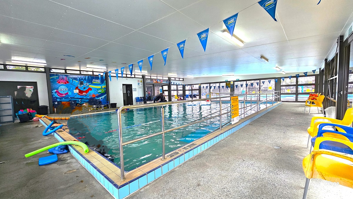 View opposite the wall with the cute swimming pool graphics. There are several pool floating toys on the poolside. A “Caution No Diving” sign is placed on the pool edge with metal handrails. Two strings of blue triangular flags across the pool are attached to the white ceiling. Blue and yellow chairs are available around the pool deck. The area is illuminated by artificial lighting and natural light from the tinted glass walls. View opposite the wall with the cute swimming pool graphics. There are several pool floating toys on the poolside. A “Caution No Diving” sign is placed on the pool edge with metal handrails. Two strings of blue triangular flags across the pool are attached to the white ceiling. Blue and yellow chairs are available around the pool deck. The area is illuminated by artificial lighting and natural light from the tinted glass walls.