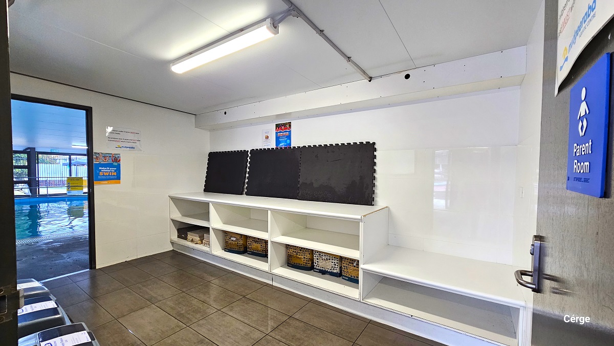 Parent room at Mudgeeraba Aquatic Centre. There are three grey garbage bins inside, a white cabinet, and three large black mats on top of the cabinet. On the lower space of the cabinet are net bags and books. The blue Parent Room sign can be found on the black door with a stainless steel door handle. The Parent room has a grey tiled floor, white walls, and ceiling. Parent room at Mudgeeraba Aquatic Centre. There are three grey garbage bins inside, a white cabinet, and three large black mats on top of the cabinet. On the lower space of the cabinet are net bags and books. The blue Parent Room sign can be found on the black door with a stainless steel door handle. The Parent room has a grey tiled floor, white walls, and ceiling.