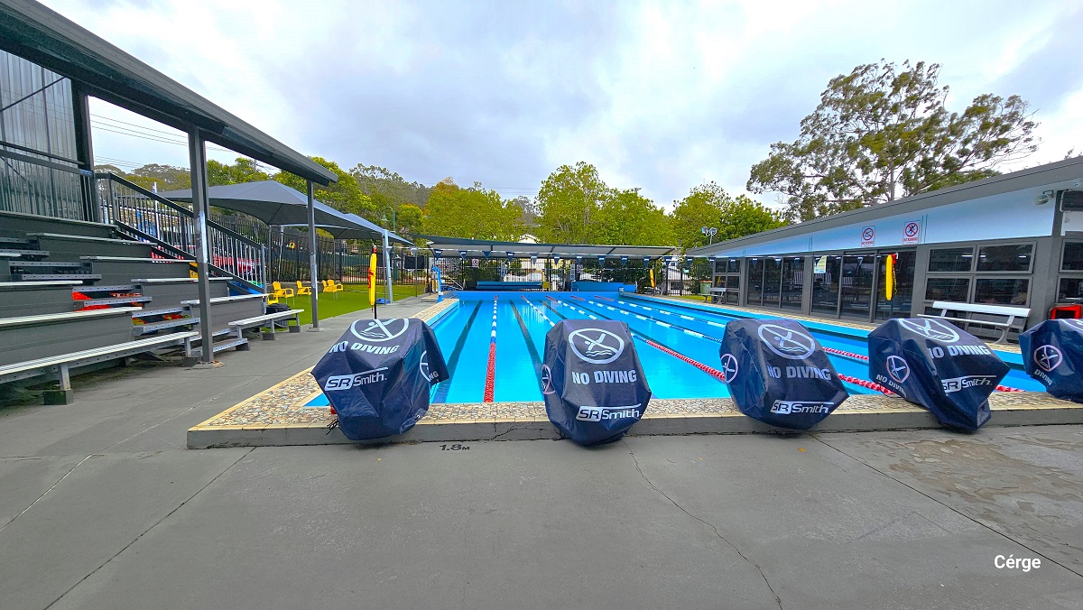 Outdoor aquatic area is directly ahead of reception with a 6-lane, 25m pool heated to a temperature setting of 26.0 - 28.0 degrees and has a depth range of 1.1m to 1.9m deep. The diving blocks are covered with a “No Diving” blue cloth. On the left side of the pool is a black-tiered seating, a handrail on the edge, covered with a black roof. Next to the tiered seating are two grey sunshades with yellow chairs. Two yellow floating devices are attached to the poles with triangular blue flags. Outdoor aquatic area is directly ahead of reception with a 6-lane, 25m pool heated to a temperature setting of 26.0 - 28.0 degrees and has a depth range of 1.1m to 1.9m deep. The diving blocks are covered with a “No Diving” blue cloth. On the left side of the pool is a black-tiered seating, a handrail on the edge, covered with a black roof. Next to the tiered seating are two grey sunshades with yellow chairs. Two yellow floating devices are attached to the poles with triangular blue flags.