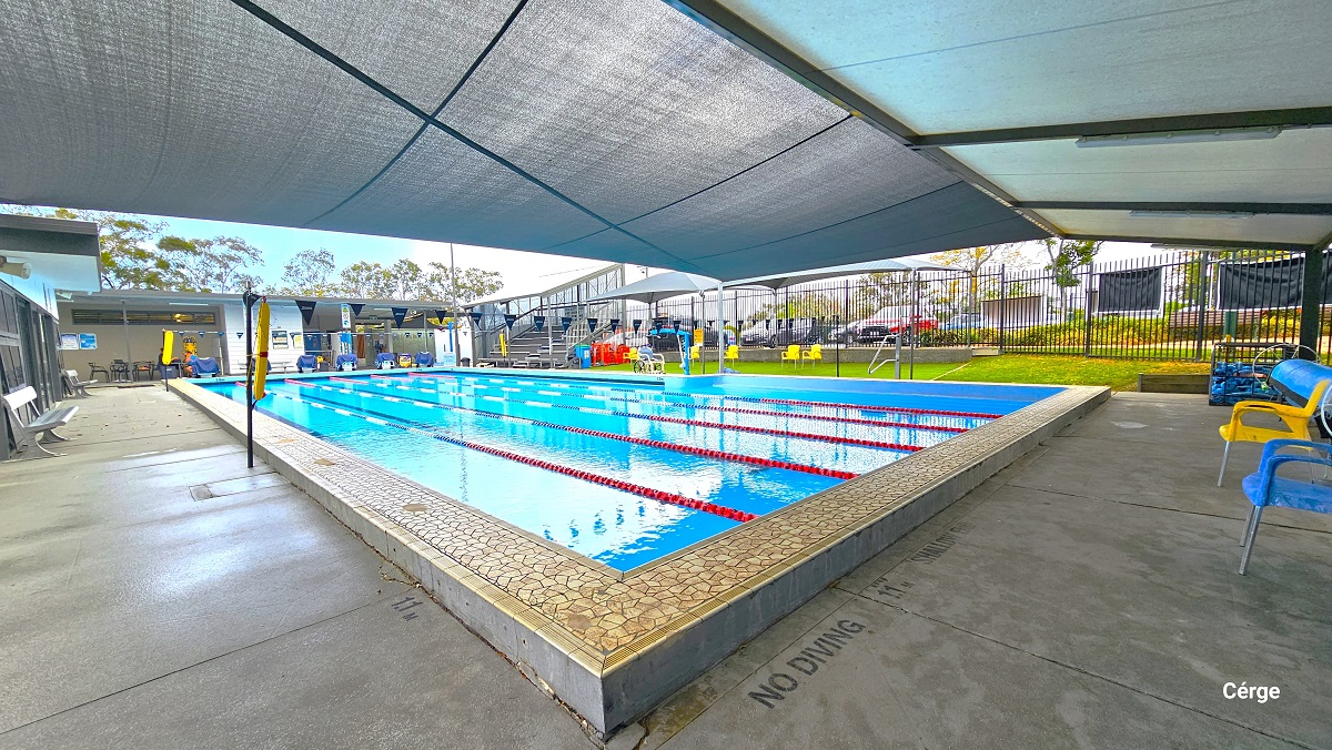 25m with 6 lanes outdoor aquatic pool at Mudgeeraba Aquatic Centre. The pool lanes are separated by red, white, and blue floating rope lanes. Two strings of triangular blue flags are tied around the opposite poles with a yellow flotation device. Seen at the back of this image are diving platforms covered with a blue cloth at the start of each pool lane, and to the right is multi-tiered seating. Yellow, blue, black, and grey plastic chairs are available all throughout the area. 25m with 6 lanes outdoor aquatic pool at Mudgeeraba Aquatic Centre. The pool lanes are separated by red, white, and blue floating rope lanes. Two strings of triangular blue flags are tied around the opposite poles with a yellow flotation device. Seen at the back of this image are diving platforms covered with a blue cloth at the start of each pool lane, and to the right is multi-tiered seating. Yellow, blue, black, and grey plastic chairs are available all throughout the area.