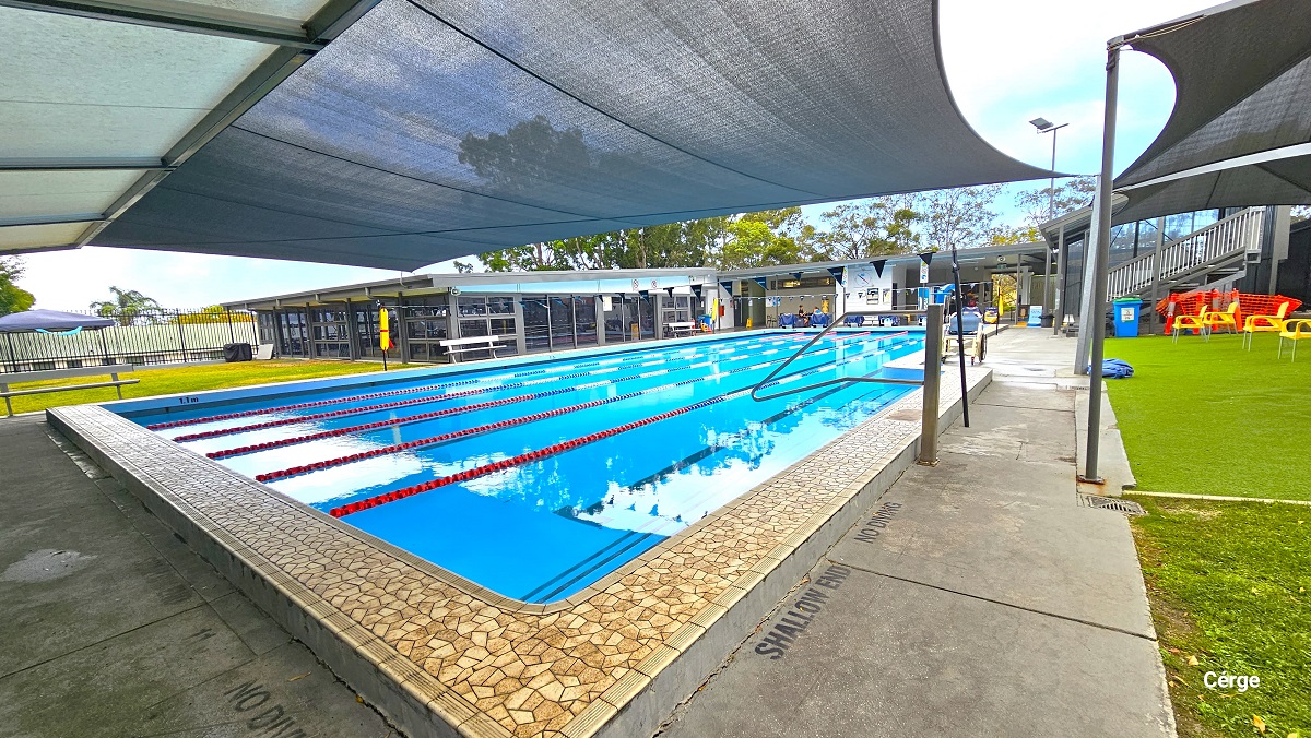 Shallow end of the 25m at Mudgeeraba Aquatic Centre, near the two black sunshades with yellow plastic chairs. It has no diving mark on the poolside. Opposite this side is a large open space with Bermuda grass, a blue tent, and a black fence. Seen above the entire facility are large trees with abundant foliage. Shallow end of the 25m at Mudgeeraba Aquatic Centre, near the two black sunshades with yellow plastic chairs. It has no diving mark on the poolside. Opposite this side is a large open space with Bermuda grass, a blue tent, and a black fence. Seen above the entire facility are large trees with abundant foliage.