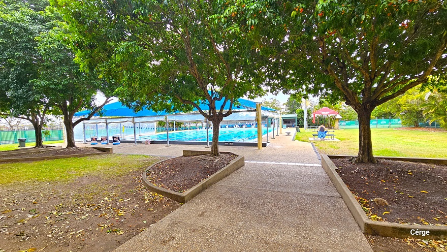 Step-free sloping pathway from the reception leads to the 25m, nine-lane lap pool at Murrumba Downs Swimming Pool. A large bright blue dome shade covers the closest section of the pool and has three signboards. A semi-circle and elongated triangular garden can be found around the area, which contains the trees that offer shade to the pathway. On the right side of the pool is a large field with a red sunshade with a table and bench. Step-free sloping pathway from the reception leads to the 25m, nine-lane lap pool at Murrumba Downs Swimming Pool. A large bright blue dome shade covers the closest section of the pool and has three signboards. A semi-circle and elongated triangular garden can be found around the area, which contains the trees that offer shade to the pathway. On the right side of the pool is a large field with a red sunshade with a table and bench.