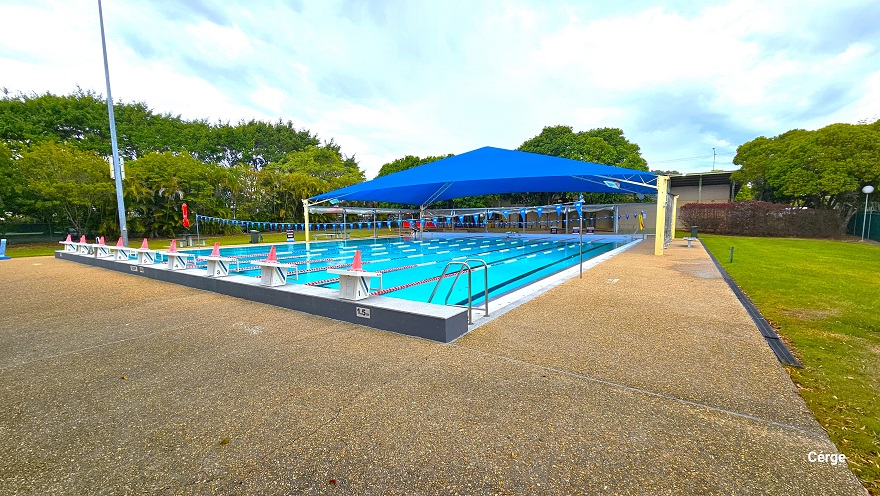 25m, nine-lane outdoor pool at Murrumba Downs Swimming Pool. The starting pool section has an elevated grey tile floor with nine white diving blocks that have orange traffic cones on top. The pool deck is brown with a rough surface to avoid slipping. The other end is partially covered by a wide blue sunshade supported by yellow metal frames. A few benches with tables are available around the pool, and strings of triangular blue flags are wrapped around steel posts on opposite pool sides. 25m, nine-lane outdoor pool at Murrumba Downs Swimming Pool. The starting pool section has an elevated grey tile floor with nine white diving blocks that have orange traffic cones on top. The pool deck is brown with a rough surface to avoid slipping. The other end is partially covered by a wide blue sunshade supported by yellow metal frames. A few benches with tables are available around the pool, and strings of triangular blue flags are wrapped around steel posts on opposite pool sides.