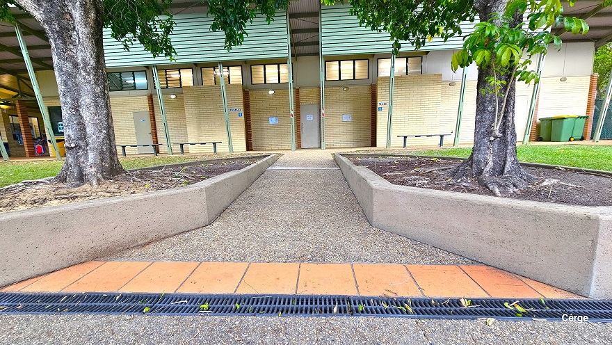 Ramp access to the accessible bathroom and changing rooms. The ramp access is between two triangular gardens in front of the bathrooms and is marked by a dark grey grating water channel and a single lane of light brown tile floor. The bathroom area has a corrugated lime green panel covering the ceiling. Grey benches are available in front. Towards the left end are large green garbage bins with black wheels. Ramp access to the accessible bathroom and changing rooms. The ramp access is between two triangular gardens in front of the bathrooms and is marked by a dark grey grating water channel and a single lane of light brown tile floor. The bathroom area has a corrugated lime green panel covering the ceiling. Grey benches are available in front. Towards the left end are large green garbage bins with black wheels.