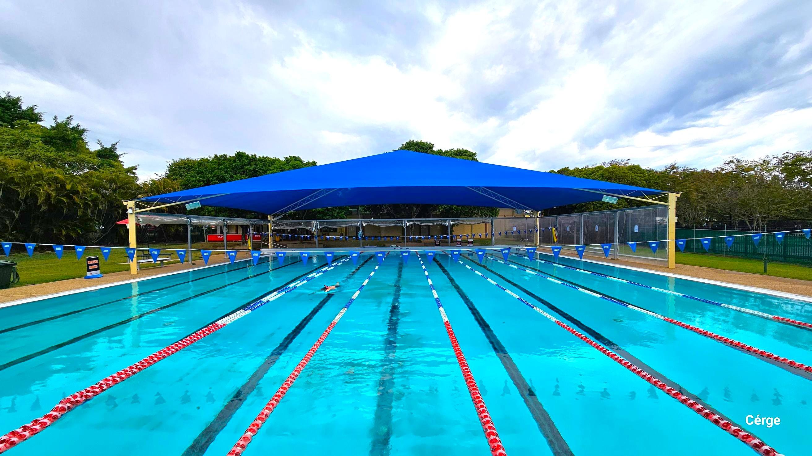 25m, nine-lane outdoor pool at Murrumba Downs Swimming Pool. The starting pool section has an elevated grey tile floor with nine white diving blocks that have orange traffic cones on top. The pool deck is brown with a rough surface to avoid slipping. The other end is partially covered by a wide blue sunshade supported by yellow metal frames. A few benches with tables are available around the pool, and strings of triangular blue flags are wrapped around steel posts on opposite pool sides. 25m, nine-lane outdoor pool at Murrumba Downs Swimming Pool. The starting pool section has an elevated grey tile floor with nine white diving blocks that have orange traffic cones on top. The pool deck is brown with a rough surface to avoid slipping. The other end is partially covered by a wide blue sunshade supported by yellow metal frames. A few benches with tables are available around the pool, and strings of triangular blue flags are wrapped around steel posts on opposite pool sides.