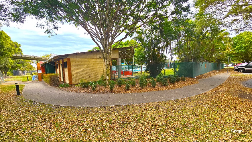 A concrete pathway with a rough surface connects the car park to a low-set cream brick building with light brown brick foundation edges. Black safety bollards can be seen on the side of the concrete pathway. Covering the landscape is dried leaf litter. Small green plants with parallel leaf venation can be seen on the right, just before the building. A concrete pathway with a rough surface connects the car park to a low-set cream brick building with light brown brick foundation edges. Black safety bollards can be seen on the side of the concrete pathway. Covering the landscape is dried leaf litter. Small green plants with parallel leaf venation can be seen on the right, just before the building.
