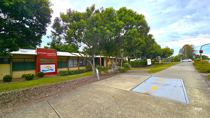 Closer look leading to another entry point closer to the main highway. Image shows several low-set small cream brick buildings with glass windows that have green outline frames and light brown brick foundation edges. Seen from this position is a large red wall cover detailing the Murrumba Community Swimming Pool on top and the logo on a white background below. Shrubs and bushes can be seen against the walls of the buildings. Closer look leading to another entry point closer to the main highway. Image shows several low-set small cream brick buildings with glass windows that have green outline frames and light brown brick foundation edges. Seen from this position is a large red wall cover detailing the Murrumba Community Swimming Pool on top and the logo on a white background below. Shrubs and bushes can be seen against the walls of the buildings.