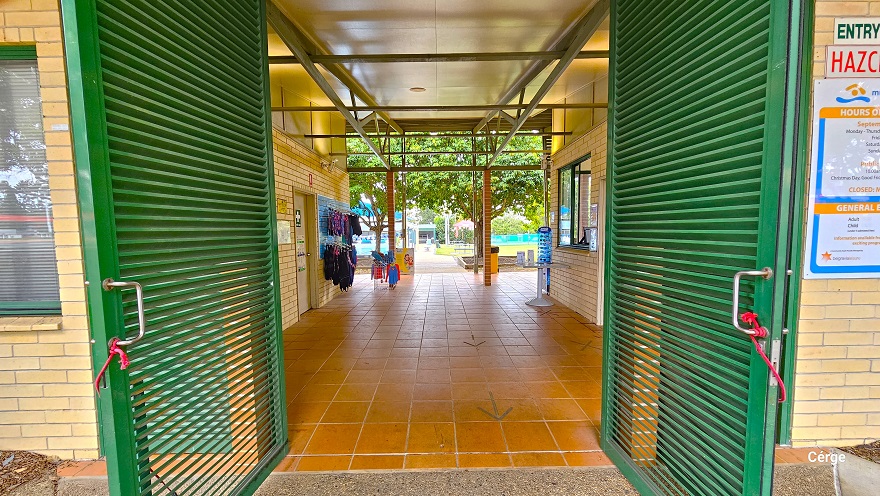 Underneath the tall entry porch with lime green metal beams and corrugated roofing of Murrumba Downs Swimming Pool. The other end of the entry porch is supported by a light brown brick foundation. Place against the left light brown brick foundation of the porch is a light blue signboard. Entrance is through green steel paneled Louvre doors right after the porch. Glass windows are noticeable on the left side. Underneath the tall entry porch with lime green metal beams and corrugated roofing of Murrumba Downs Swimming Pool. The other end of the entry porch is supported by a light brown brick foundation. Place against the left light brown brick foundation of the porch is a light blue signboard. Entrance is through green steel paneled Louvre doors right after the porch. Glass windows are noticeable on the left side.
