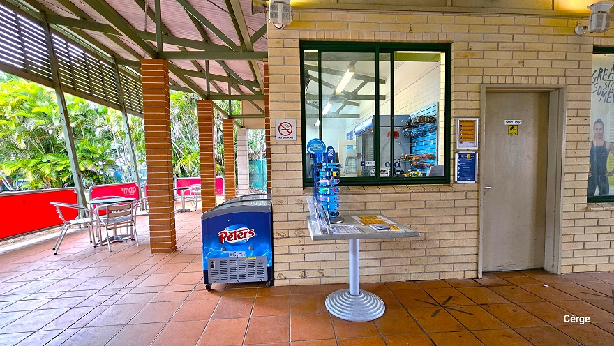 Reception and retail at Murrumba Downs Swimming. On the right side of the entrance is the ‘Staff Only’ brown door with a yellow CCTV sign. Next to the door is a glass window with green metal frames. In front of the window is a grey table with several small adverts and swimming goggles for kids placed on a blue display. A ‘No Smoking’ sign is on the left side of the window. In front of the reception is a blue fridge and stainless steel chairs and tables. Reception and retail at Murrumba Downs Swimming. On the right side of the entrance is the ‘Staff Only’ brown door with a yellow CCTV sign. Next to the door is a glass window with green metal frames. In front of the window is a grey table with several small adverts and swimming goggles for kids placed on a blue display. A ‘No Smoking’ sign is on the left side of the window. In front of the reception is a blue fridge and stainless steel chairs and tables.