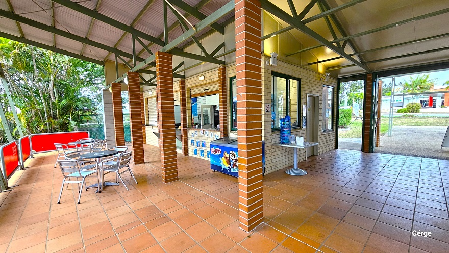 Side angle of the reception and its seating area in front. Separating the reception from the pool are low red barricades with grey steel frames. On the cream brick wall below the reception countertop is ‘GO SWIM’ and several blue graphics. The building has light brown brick foundations, narrow green metal roof beams, and corrugated roofing. The reception area has cream roll-down shutters with light brown accents. Side angle of the reception and its seating area in front. Separating the reception from the pool are low red barricades with grey steel frames. On the cream brick wall below the reception countertop is ‘GO SWIM’ and several blue graphics. The building has light brown brick foundations, narrow green metal roof beams, and corrugated roofing. The reception area has cream roll-down shutters with light brown accents.