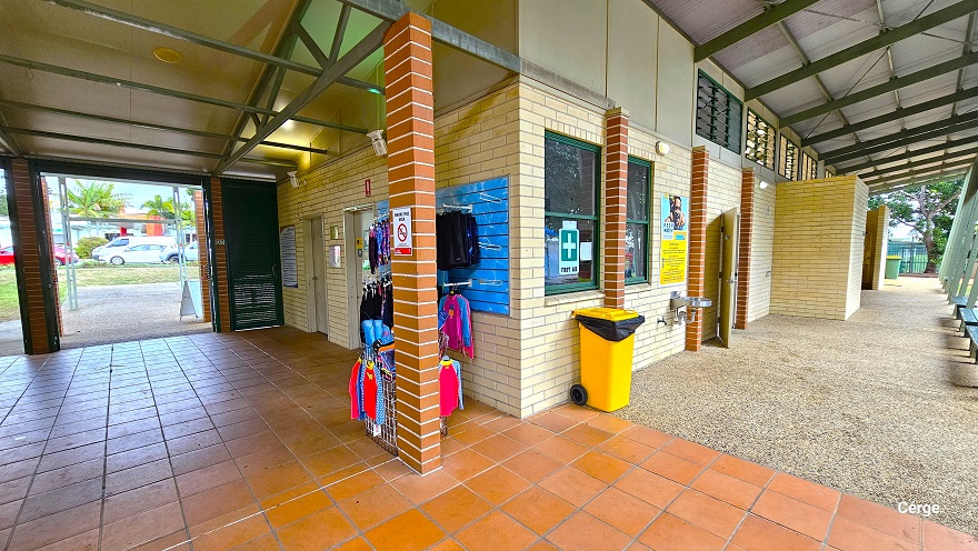 Found opposite the reception at Murrumba Downs Swimming Pool are two white doors accessible along the corridor right after passing the green steel panel Louvre doors. Next to the second white door is the kid's swimwear display. There is a small white ‘No Smoking’ sign on the light brown brick foundation. In front of the glass window with a green cross is a yellow garbage bin with wheels. Open windows near the ceiling provide ventilation for the male and female change rooms. Found opposite the reception at Murrumba Downs Swimming Pool are two white doors accessible along the corridor right after passing the green steel panel Louvre doors. Next to the second white door is the kid's swimwear display. There is a small white ‘No Smoking’ sign on the light brown brick foundation. In front of the glass window with a green cross is a yellow garbage bin with wheels. Open windows near the ceiling provide ventilation for the male and female change rooms.