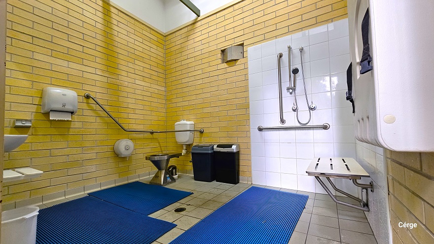 Inside the accessible bathroom at Murrumba Downs Swimming Pool. Near the door is a white adult change table with blue straps. Next to it is the shower section which is highlighted by the white tile wall that contains the shower head, grab bars, and a wall-mounted white shower seat. On the grey tile floor are blue waterproof mats. At the back is the toilet, two black garbage bins, and a grab bar. On the left wall is a white tissue dispenser, a wash basin, and a white pail. Inside the accessible bathroom at Murrumba Downs Swimming Pool. Near the door is a white adult change table with blue straps. Next to it is the shower section which is highlighted by the white tile wall that contains the shower head, grab bars, and a wall-mounted white shower seat. On the grey tile floor are blue waterproof mats. At the back is the toilet, two black garbage bins, and a grab bar. On the left wall is a white tissue dispenser, a wash basin, and a white pail.