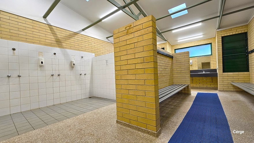 Shower section at Murrumba Downs Swimming Pool has a white tile wall and grey tile floor. A light brown brick wall separates the shower from the waiting area and has wall wall-mounted grey bench and a granite tile floor with a long blue waterproof floor mat at the center. There is a dark wash area towards the exit with a wash basin and a liquid hand soap dispenser. The shower room has a white ceiling with narrow green metal roof beams. Shower section at Murrumba Downs Swimming Pool has a white tile wall and grey tile floor. A light brown brick wall separates the shower from the waiting area and has wall wall-mounted grey bench and a granite tile floor with a long blue waterproof floor mat at the center. There is a dark wash area towards the exit with a wash basin and a liquid hand soap dispenser. The shower room has a white ceiling with narrow green metal roof beams.