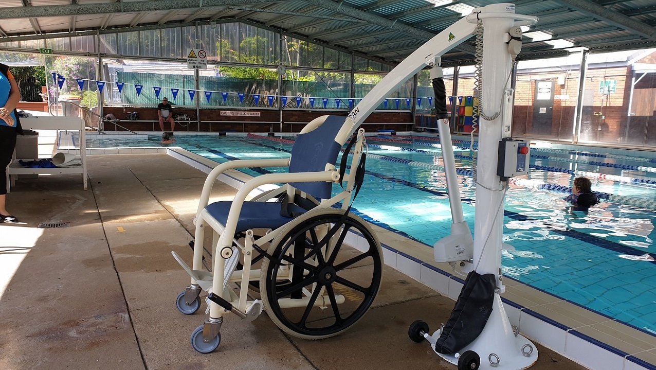 Entry to the 25m Indoor pool includes a pool hoist.  A white framed aquatic chair sits attached to a pool hoist to showcase inclusion for mobility disability.