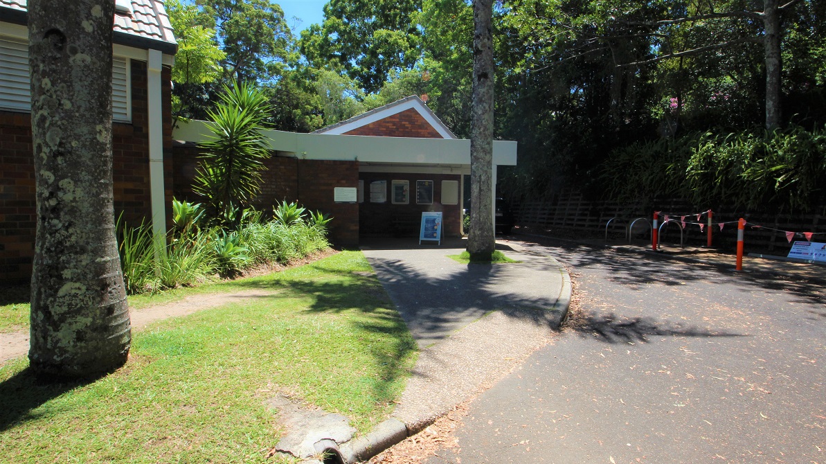 Parking in the accessible car spaces directly in front of the Nambour Aquatic Centre shows a step free entrance toward the low set brown brick building.  The venue is set amongst dense foliage creating significand shading.