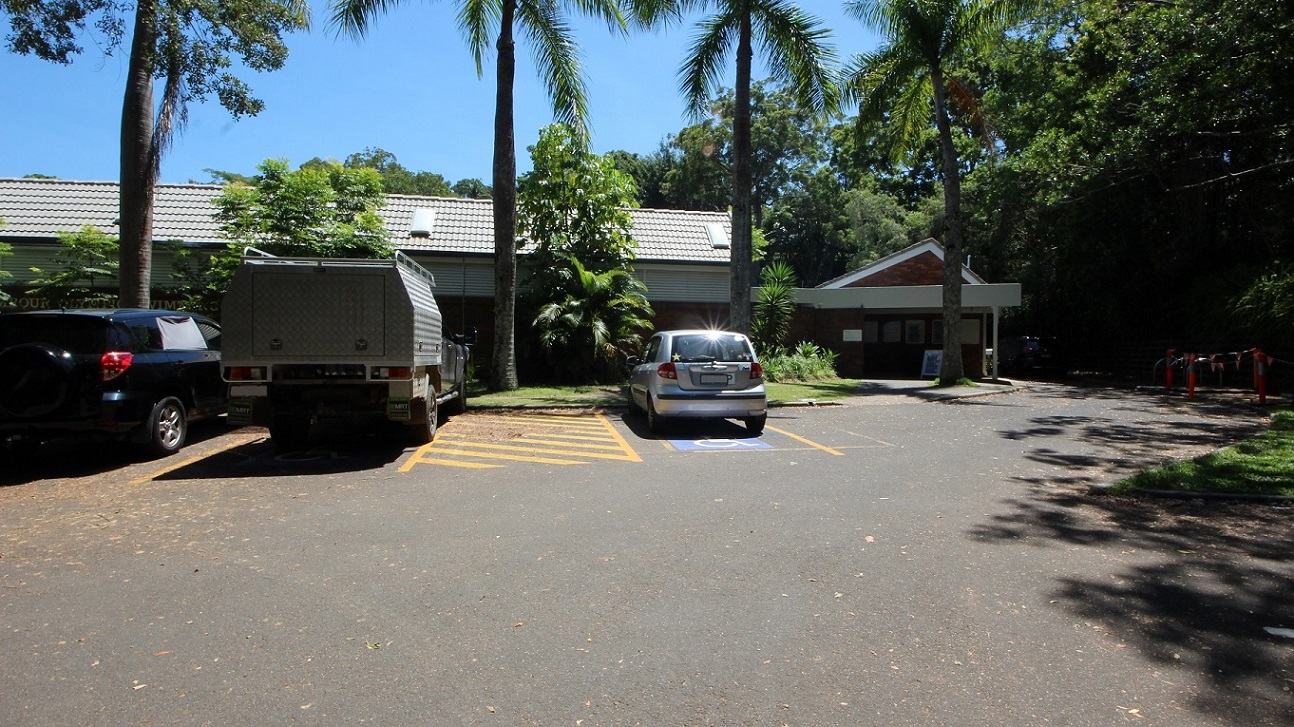 Parking in the accessible car spaces directly in front of the Nambour Aquatic Centre shows a step free entrance toward the low set brown brick building.  The venue is set amongst dense foliage creating significand shading.