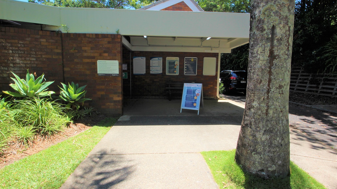 The entrance to the Nambour Aquatic Centre is a step free entrance toward the low set brown brick building.  The venue is set amongst dense foliage creating significant shading.