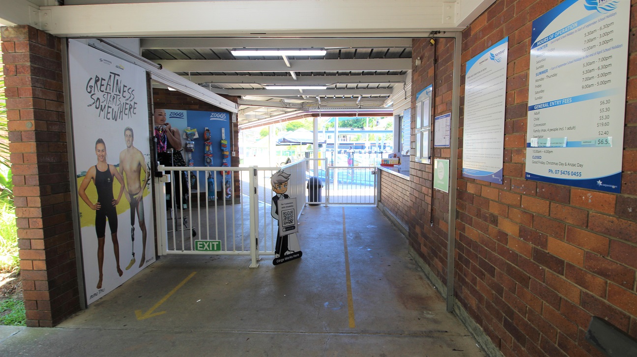 Entrance to the Nambour Aquatic Centre is a passageway design attaching to the reception counter.  Entry is via the right and exit via the left.  The concrete path and brown brick walls are combined with a white gate to manage the flow of customers.