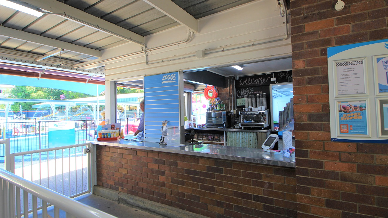 Entrance to the Nambour Aquatic Centre is a passageway design attaching to the reception counter.  Entry is via the right and exit via the left.  The concrete path and brown brick walls are combined with a white gate to manage the flow of customers.