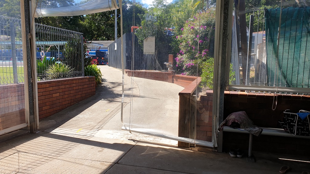 At the bottom of the step free ramp to the 25m indoor pool looking back up toward the venue reception.  In the image is also the concrete ramp to access the pool, small brown brick retaining walls and plastic walls of the indoor pool.