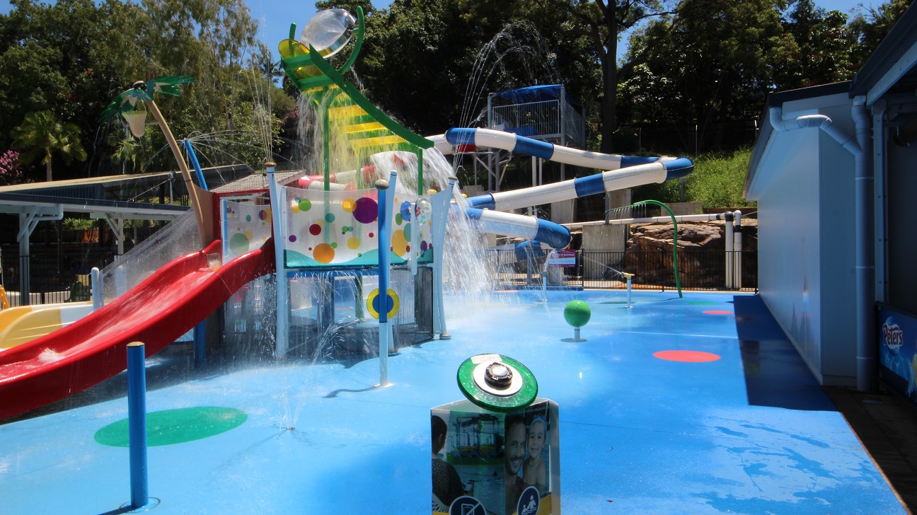The Nambour Aquatic Centre Splash Park.  The aqua blue flooring is contrasted with the red slides and yellow and green equipment.  A lot of water is splashing around the equipment. A standard pool fence bounds the splash park from the remainder of the Nambour Aquatic Centre.