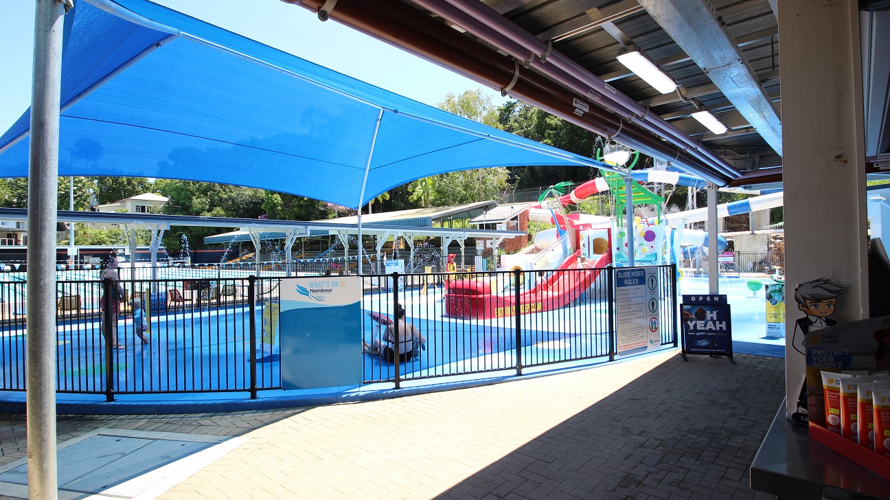The Nambour Aquatic Centre Splash Park.  The aqua blue flooring is contrasted with the red slides and yellow and green equipment.  A lot of water is splashing around the equipment.