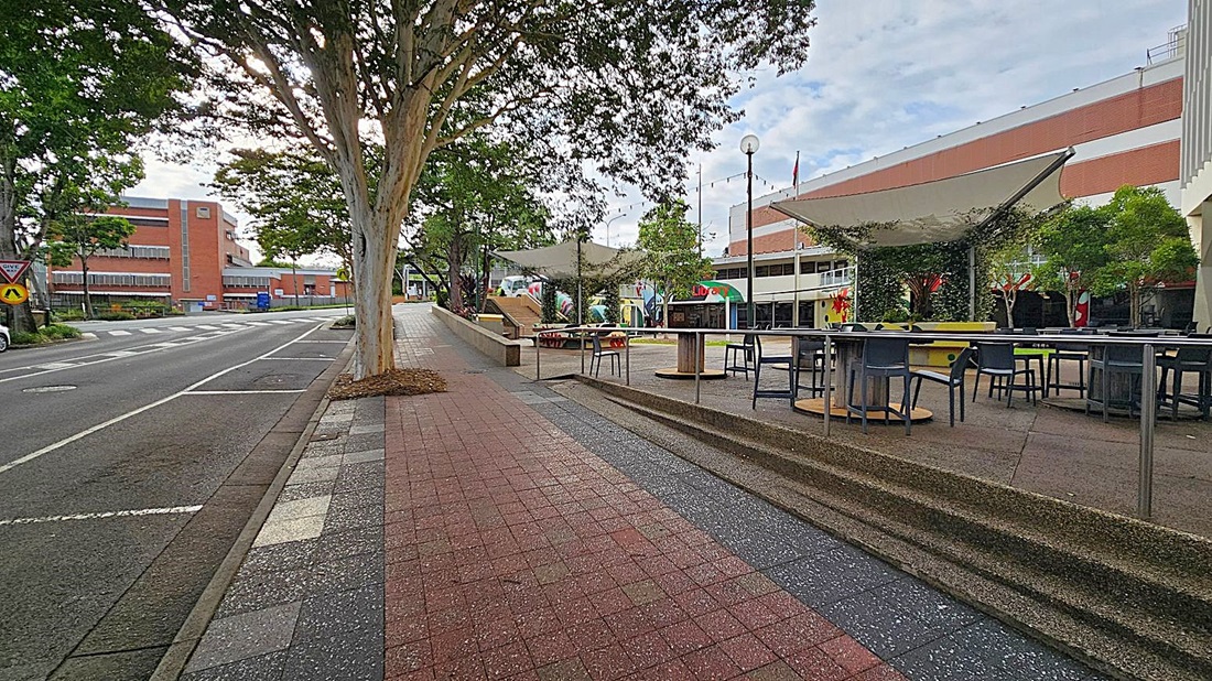 A street scene with a footpath on the left featuring a metal railing and a grey chair. Trees line the path, providing shade. On the right, a road with parked cars and a row of buildings, including a small structure with a red awning. Several bins are lined up against a building. The pavement is a mix of red and grey tiles, and a large tree trunk is visible in the foreground. A street scene with a footpath on the left featuring a metal railing and a grey chair. Trees line the path, providing shade. On the right, a road with parked cars and a row of buildings, including a small structure with a red awning. Several bins are lined up against a building. The pavement is a mix of red and grey tiles, and a large tree trunk is visible in the foreground.