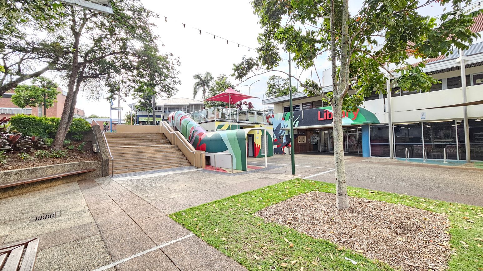 A public space with a library on the right, featuring a green and red awning. In the centre, a colourful, curved structure with a red umbrella on top leads to a staircase. On the left, there are trees and shrubs with a bench along a raised garden bed. The ground is paved with concrete, and a small patch of grass with a tree is in the foreground. Overhead, string lights are visible. A public space with a library on the right, featuring a green and red awning. In the centre, a colourful, curved structure with a red umbrella on top leads to a staircase. On the left, there are trees and shrubs with a bench along a raised garden bed. The ground is paved with concrete, and a small patch of grass with a tree is in the foreground. Overhead, string lights are visible.