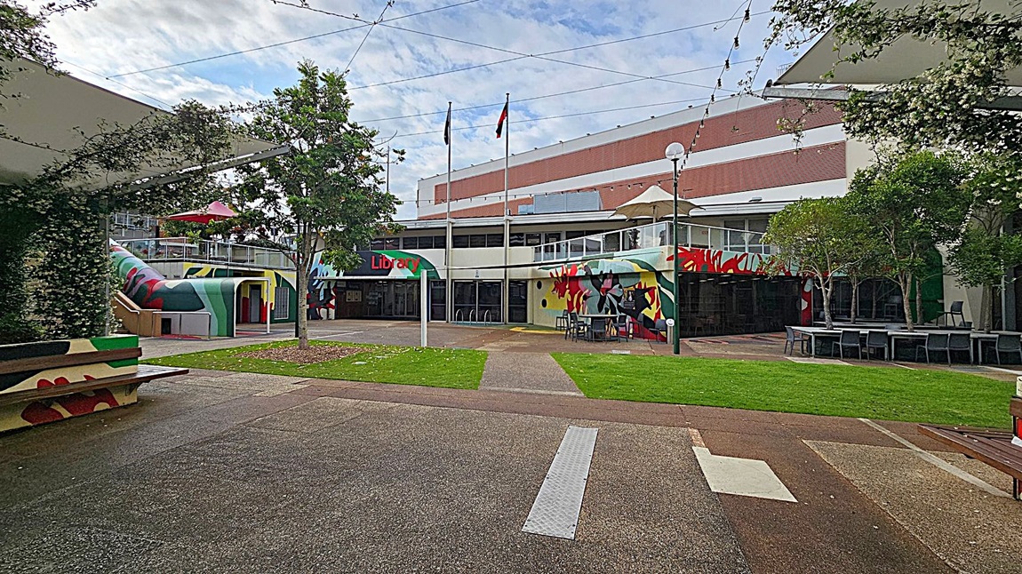 A courtyard with a library in the background. The library has colourful murals on its walls and a sign reading "Library" above the entrance. Two flagpoles stand in front of the building. On the left, a covered area with greenery and a bench. On the right, tables and chairs under trees. The ground is a mix of grass and paved paths, and string lights are strung overhead. A courtyard with a library in the background. The library has colourful murals on its walls and a sign reading "Library" above the entrance. Two flagpoles stand in front of the building. On the left, a covered area with greenery and a bench. On the right, tables and chairs under trees. The ground is a mix of grass and paved paths, and string lights are strung overhead.