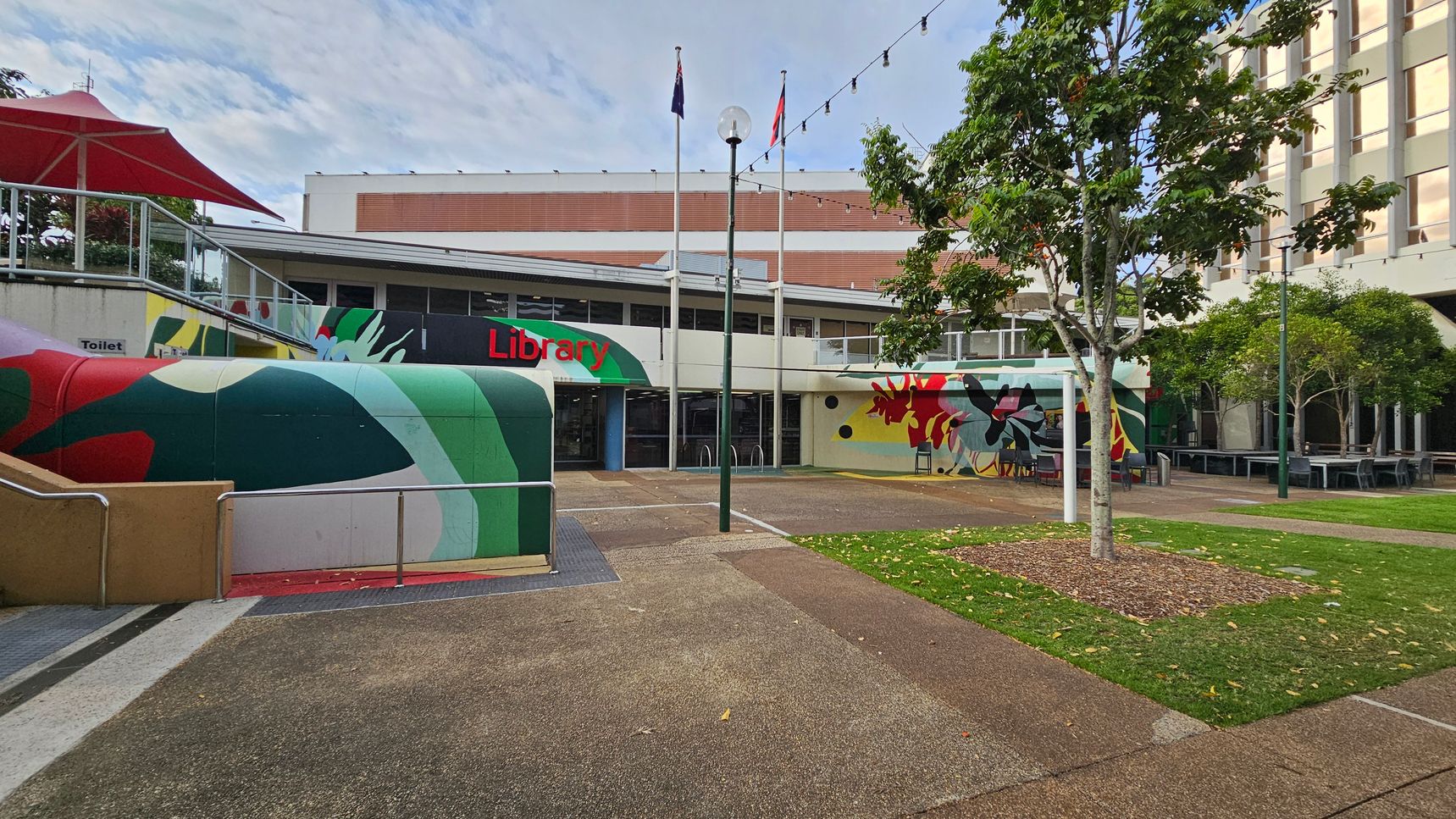 A library building with a colourful mural on the left side, featuring abstract shapes in green, red, and yellow. The entrance is in the centre, with the word "Library" in red. Three flagpoles stand in front, and a string of lights is strung above. On the right, there are trees and outdoor seating. The ground is a mix of concrete paths and grass patches. A red umbrella is visible on the left. A library building with a colourful mural on the left side, featuring abstract shapes in green, red, and yellow. The entrance is in the centre, with the word "Library" in red. Three flagpoles stand in front, and a string of lights is strung above. On the right, there are trees and outdoor seating. The ground is a mix of concrete paths and grass patches. A red umbrella is visible on the left.