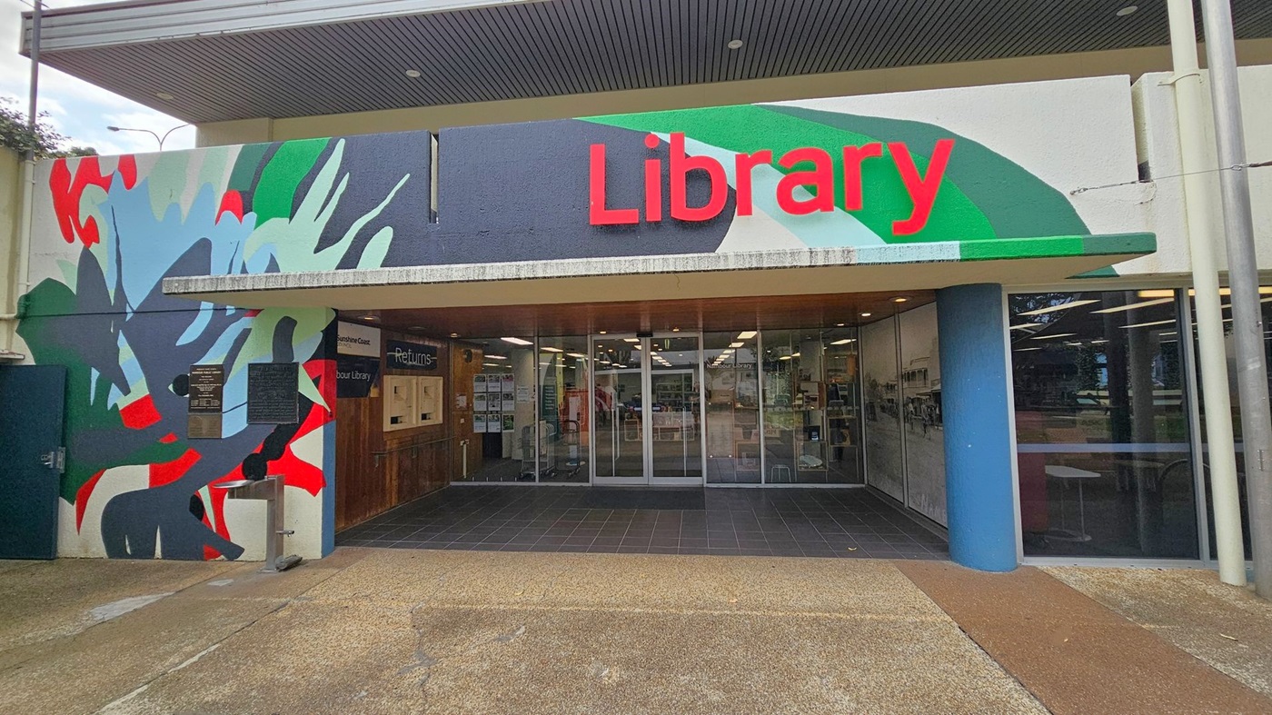 Entrance to a library with a colourful mural on the left featuring abstract shapes in red, green, and blue. The word "Library" is prominently displayed in red on the right. Glass doors lead into the building, with a returns section visible on the left. The floor outside is a mix of concrete and tiles, and the ceiling is dark with recessed lighting. Entrance to a library with a colourful mural on the left featuring abstract shapes in red, green, and blue. The word "Library" is prominently displayed in red on the right. Glass doors lead into the building, with a returns section visible on the left. The floor outside is a mix of concrete and tiles, and the ceiling is dark with recessed lighting.
