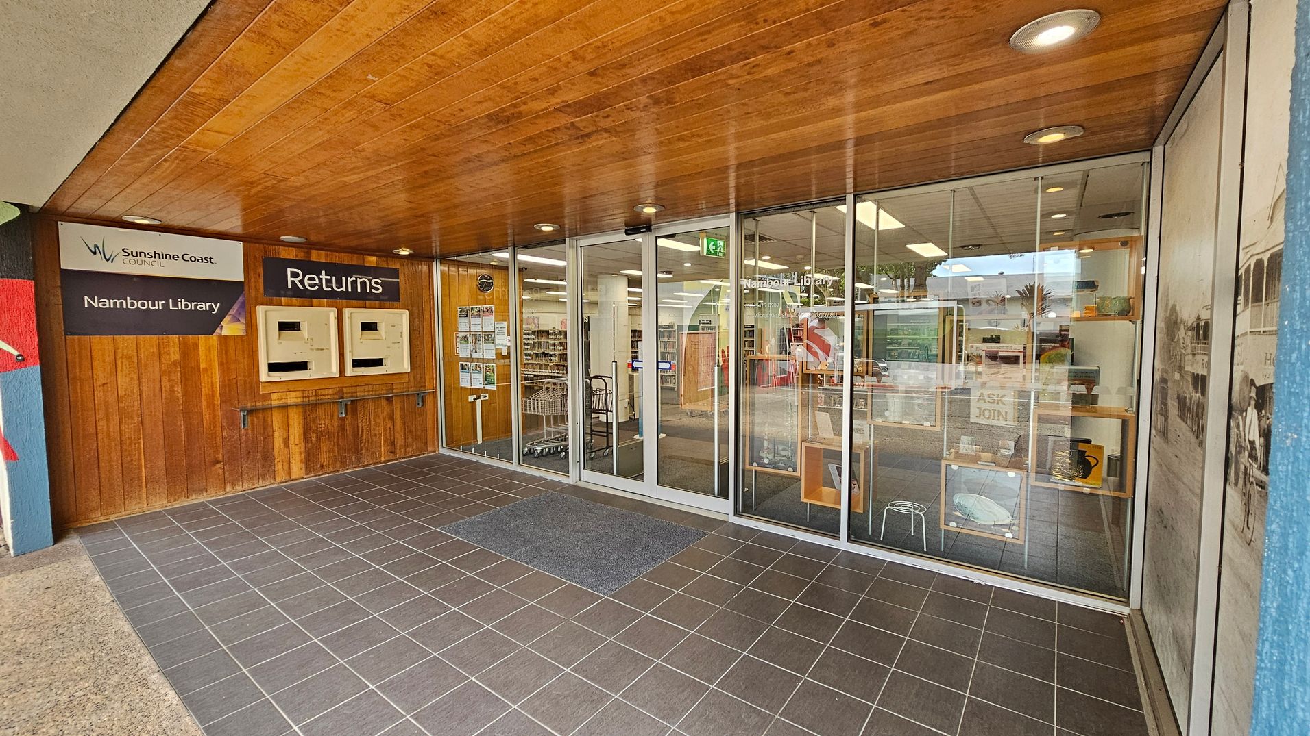 Entrance to Nambour Library with a wooden ceiling and tiled floor. On the left, there are two return slots under a "Returns" sign, and a "Sunshine Coast Council Nambour Library" sign. The glass doors in the centre lead into the library, revealing bookshelves and a cart inside. Posters and notices are displayed on the glass. On the right, a large window shows a historical photo and some library displays. Entrance to Nambour Library with a wooden ceiling and tiled floor. On the left, there are two return slots under a "Returns" sign, and a "Sunshine Coast Council Nambour Library" sign. The glass doors in the centre lead into the library, revealing bookshelves and a cart inside. Posters and notices are displayed on the glass. On the right, a large window shows a historical photo and some library displays.
