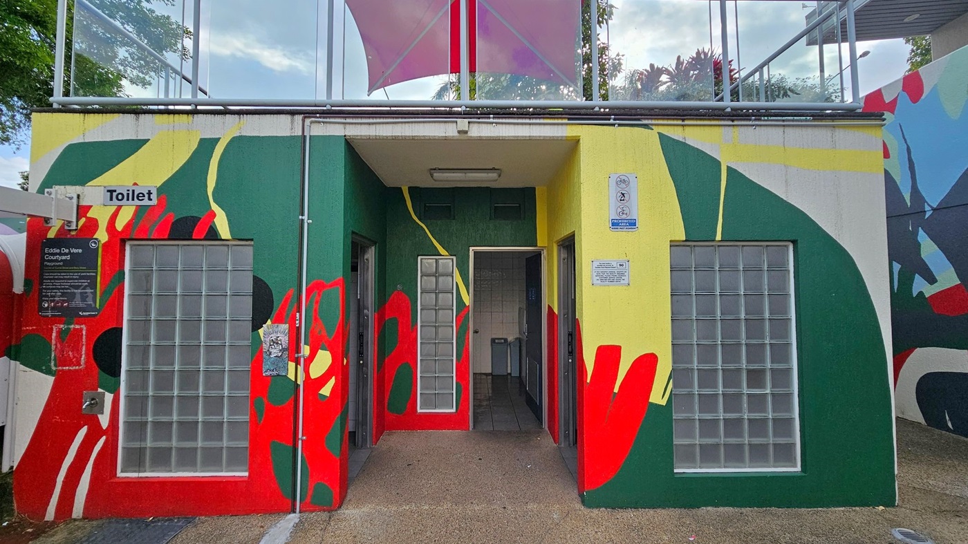 A public toilet building with a colourful mural featuring green, red, yellow, and black abstract shapes. The entrance is in the centre, flanked by glass block windows. A sign above the left side reads "Toilet," and there's a plaque for "Eddie De Vere Courtyard" below it. The ground is paved, and a pink canopy is visible above. Trees and a railing are in the background. A public toilet building with a colourful mural featuring green, red, yellow, and black abstract shapes. The entrance is in the centre, flanked by glass block windows. A sign above the left side reads "Toilet," and there's a plaque for "Eddie De Vere Courtyard" below it. The ground is paved, and a pink canopy is visible above. Trees and a railing are in the background.