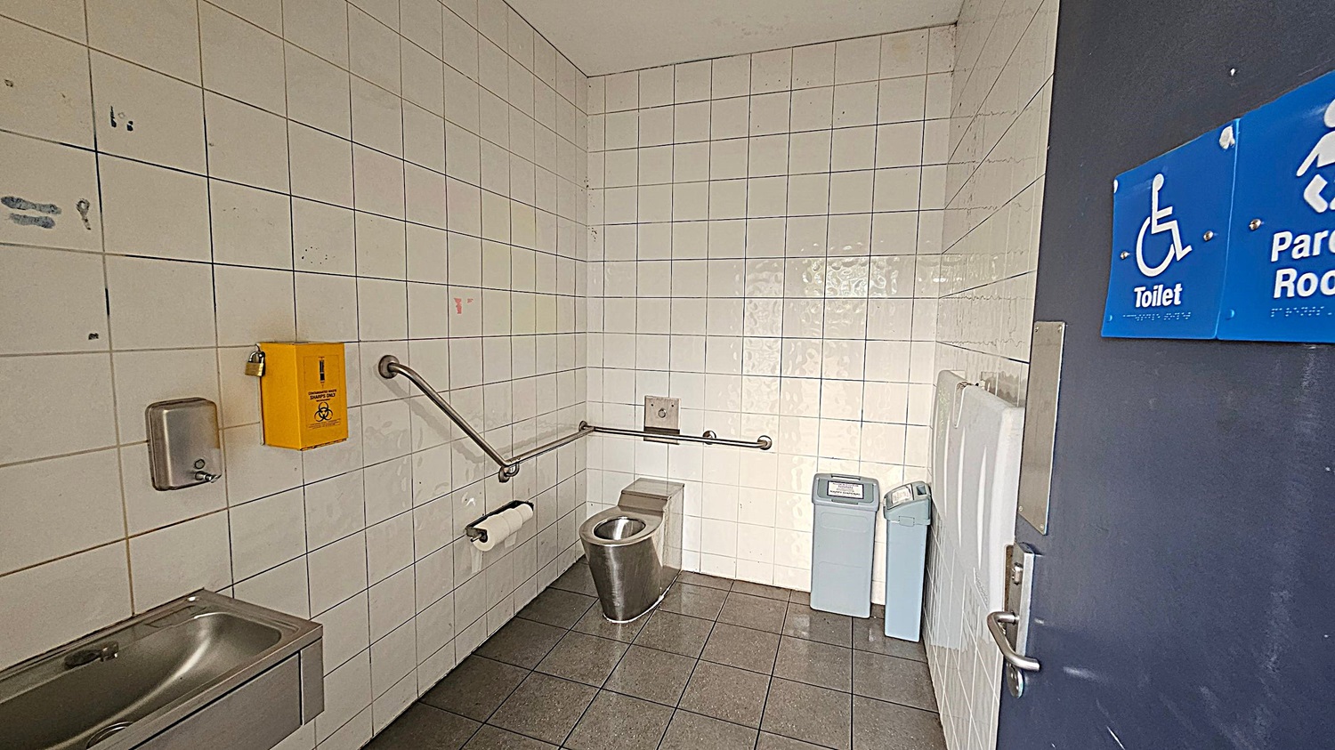 A public accessible toilet with white tiled walls and a grey tiled floor. On the left, there's a stainless steel sink with a soap dispenser above it and a yellow biohazard disposal box. A metal grab rail runs along the wall, with a toilet roll holder attached. In the centre, a stainless steel toilet is positioned against the wall. On the right, there are two bins and a baby changing station. The door has a blue sign indicating it's a toilet and parent room. A public accessible toilet with white tiled walls and a grey tiled floor. On the left, there's a stainless steel sink with a soap dispenser above it and a yellow biohazard disposal box. A metal grab rail runs along the wall, with a toilet roll holder attached. In the centre, a stainless steel toilet is positioned against the wall. On the right, there are two bins and a baby changing station. The door has a blue sign indicating it's a toilet and parent room.