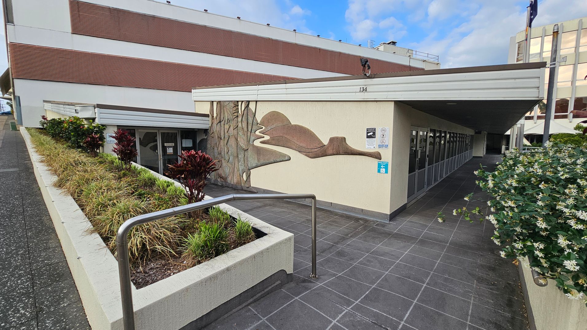 A building with a mural on the wall, featuring abstract shapes in earthy tones. On the left, there's a sloped path with a metal railing and a planter box filled with grass and red-leaved plants. The right side shows a row of glass doors under a flat roof. In the foreground, a bush with white flowers is visible. The ground is paved with dark tiles, and the building has a mix of white and brown exterior panels. A building with a mural on the wall, featuring abstract shapes in earthy tones. On the left, there's a sloped path with a metal railing and a planter box filled with grass and red-leaved plants. The right side shows a row of glass doors under a flat roof. In the foreground, a bush with white flowers is visible. The ground is paved with dark tiles, and the building has a mix of white and brown exterior panels.