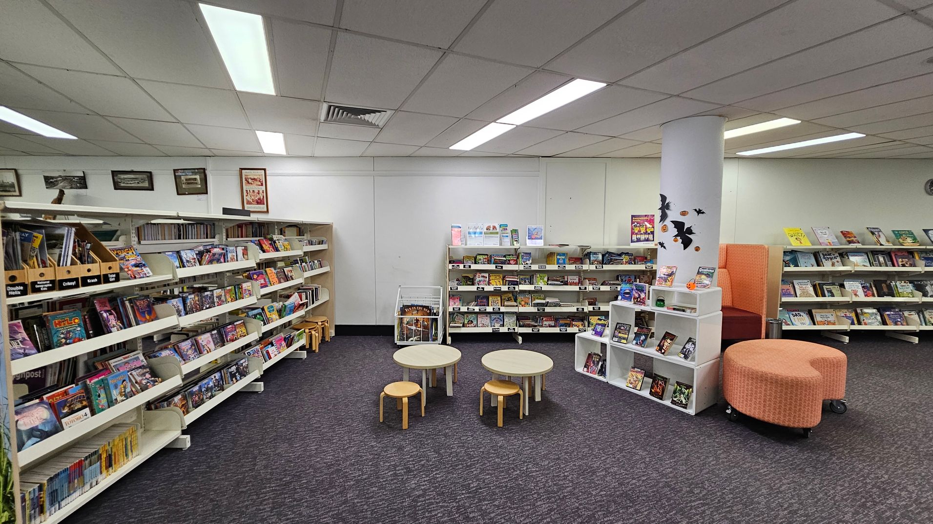 A library interior with bookshelves lining the left and right walls, filled with various books and magazines. In the centre, there are small round tables with wooden stools. A white pillar decorated with black bat cutouts stands near an orange cushioned seating area. The floor is carpeted in dark grey, and the ceiling has fluorescent lights and a grid pattern. A library interior with bookshelves lining the left and right walls, filled with various books and magazines. In the centre, there are small round tables with wooden stools. A white pillar decorated with black bat cutouts stands near an orange cushioned seating area. The floor is carpeted in dark grey, and the ceiling has fluorescent lights and a grid pattern.