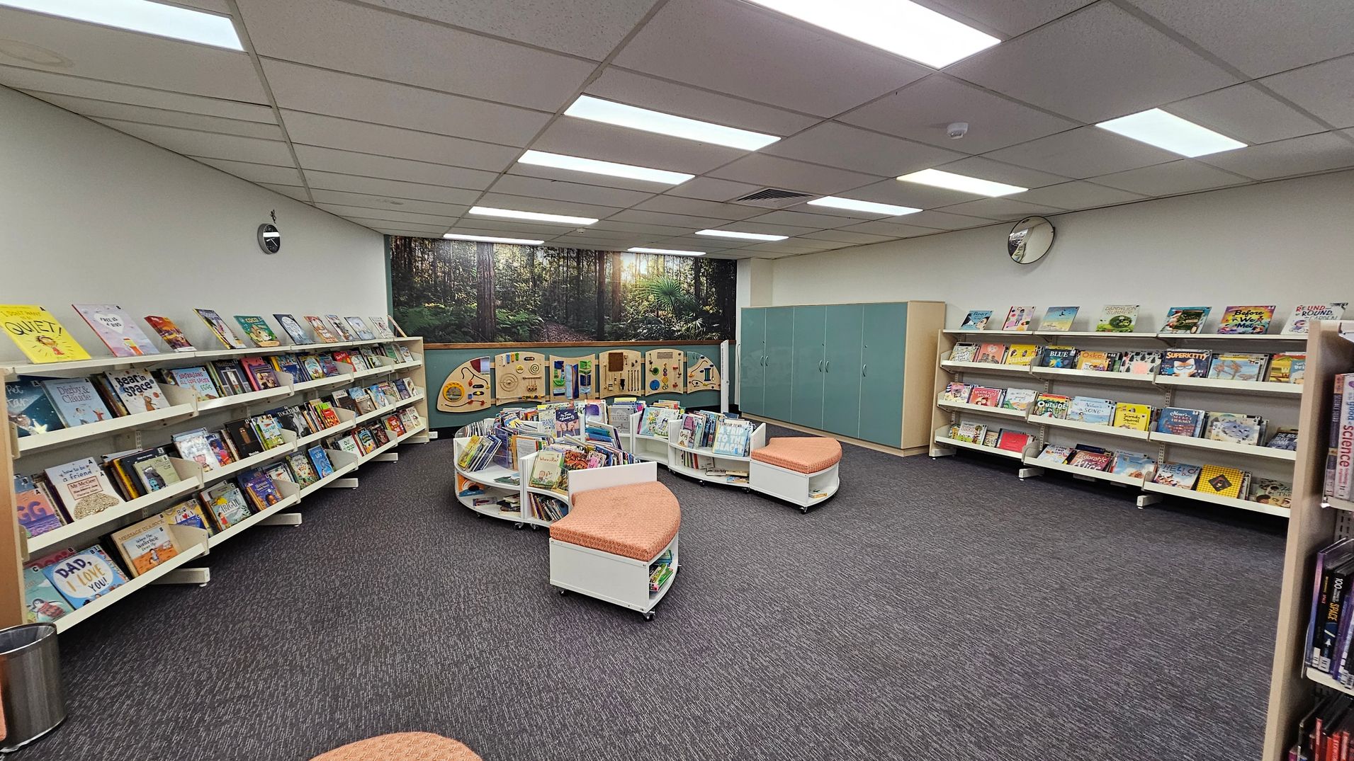 A library room with bookshelves lining the left and right walls, filled with children's books. In the centre, there are curved shelves with more books and cushioned seating. The back wall features a mural of a forest scene and green cabinets. The ceiling has fluorescent lights, and the floor is carpeted in dark grey. A round clock is mounted on the right wall. A library room with bookshelves lining the left and right walls, filled with children's books. In the centre, there are curved shelves with more books and cushioned seating. The back wall features a mural of a forest scene and green cabinets. The ceiling has fluorescent lights, and the floor is carpeted in dark grey. A round clock is mounted on the right wall.