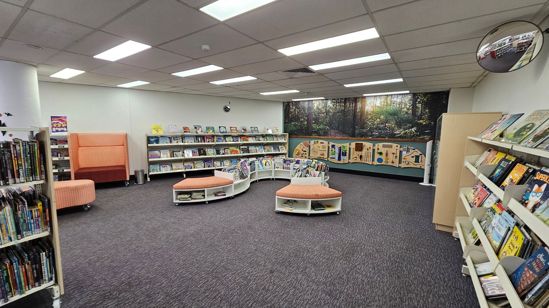 A library interior with grey carpeted flooring and a ceiling with fluorescent lights. On the left, there are shelves filled with books and DVDs. In the centre, there are curved bookshelves with orange cushioned seating. The back wall features a large mural of a forest scene and interactive panels. On the right, more bookshelves display children's books. A circular mirror is mounted on the upper right wall. A library interior with grey carpeted flooring and a ceiling with fluorescent lights. On the left, there are shelves filled with books and DVDs. In the centre, there are curved bookshelves with orange cushioned seating. The back wall features a large mural of a forest scene and interactive panels. On the right, more bookshelves display children's books. A circular mirror is mounted on the upper right wall.