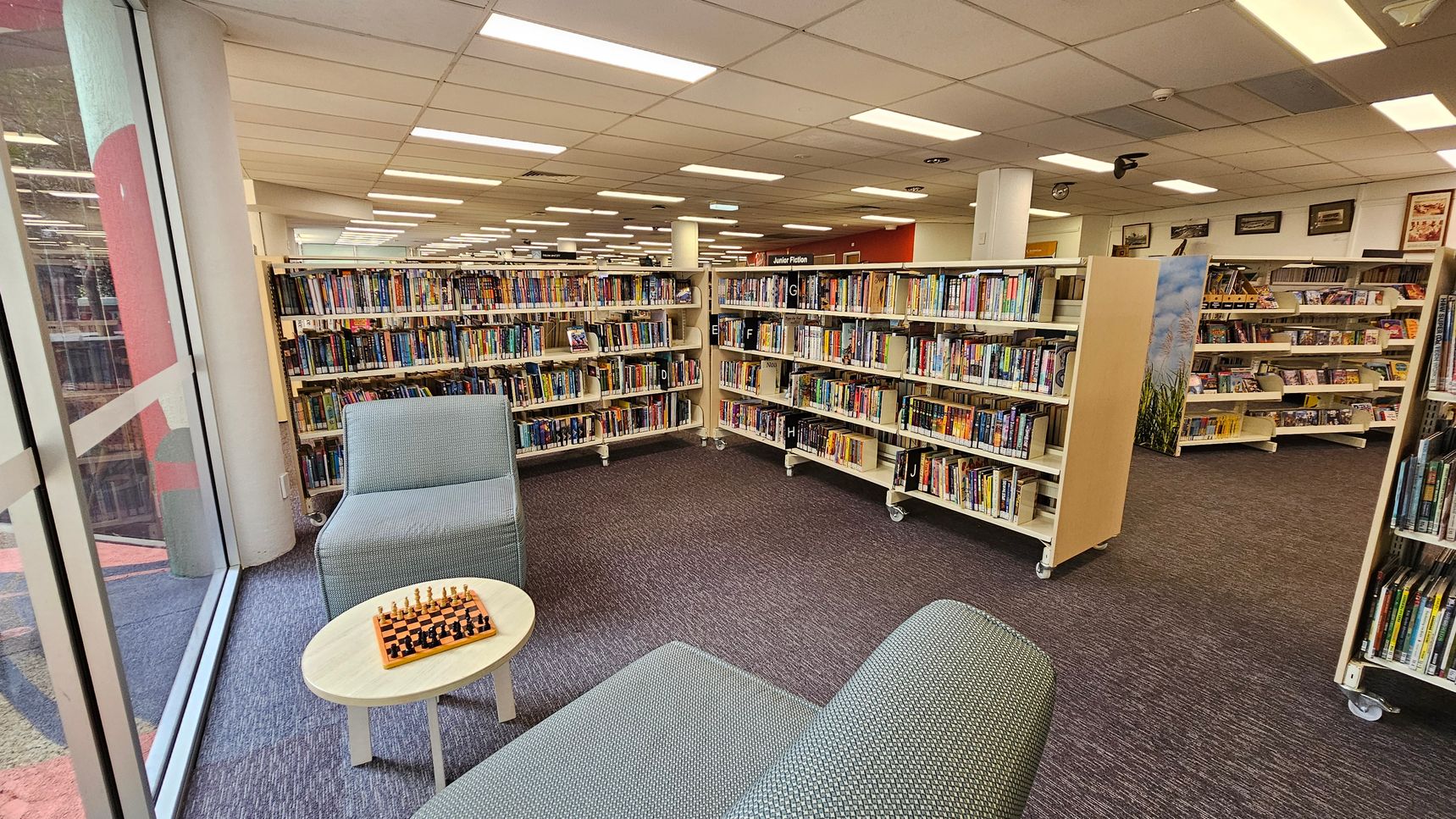 A library interior with bookshelves filled with books on the right and in the background. In the foreground, there are two grey upholstered chairs and a small round table with a chessboard on it. The floor is carpeted in a dark grey colour. Large windows on the left allow natural light to enter. The ceiling is lined with fluorescent lights. A library interior with bookshelves filled with books on the right and in the background. In the foreground, there are two grey upholstered chairs and a small round table with a chessboard on it. The floor is carpeted in a dark grey colour. Large windows on the left allow natural light to enter. The ceiling is lined with fluorescent lights.