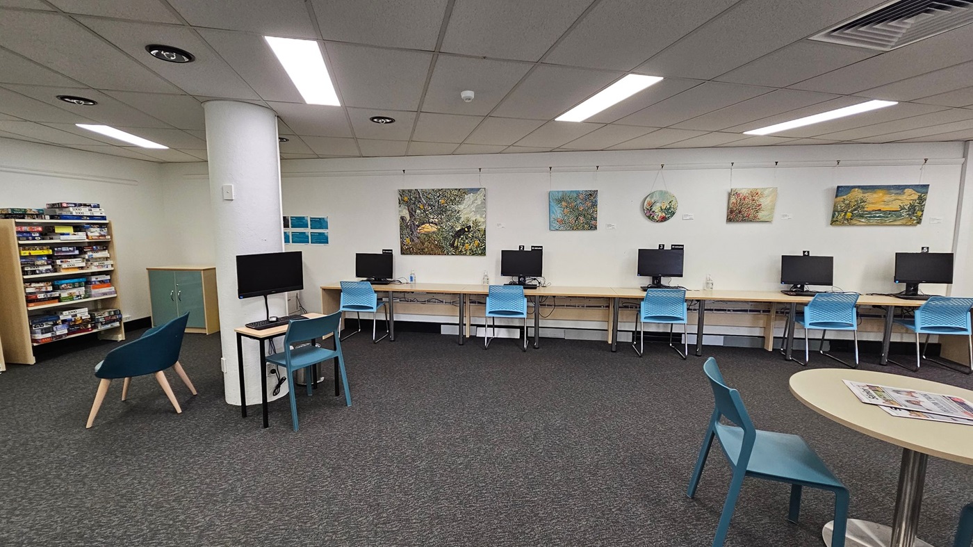 A library interior with a grey carpeted floor and a white panelled ceiling with fluorescent lights. On the left, there are bookshelves labelled "Audiobooks" filled with items. A teal chair is positioned near a small table. On the right, there are computer stations with blue chairs and monitors on wooden desks. A large painting hangs above the computers. A white column stands in the centre of the room. A library interior with a grey carpeted floor and a white panelled ceiling with fluorescent lights. On the left, there are bookshelves labelled "Audiobooks" filled with items. A teal chair is positioned near a small table. On the right, there are computer stations with blue chairs and monitors on wooden desks. A large painting hangs above the computers. A white column stands in the centre of the room.