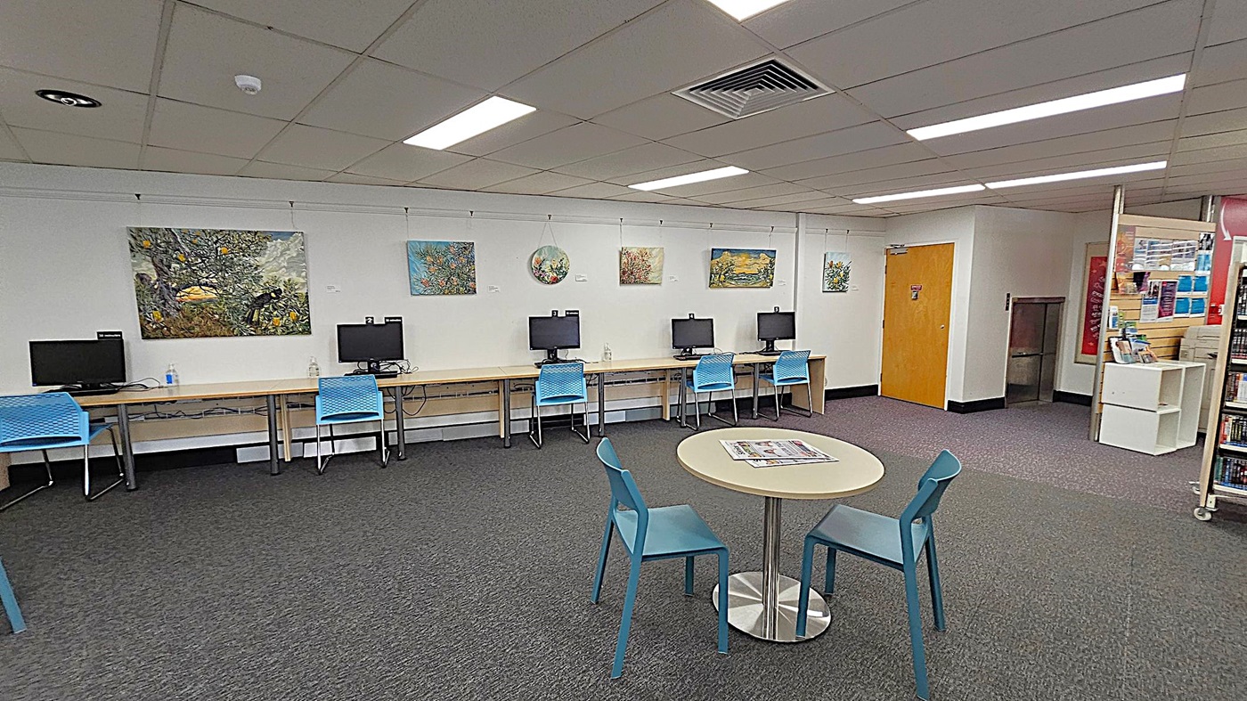 A room with a row of computers on desks against the left wall, each accompanied by a blue chair. Above the computers, several colourful paintings are displayed. In the foreground, a round table with two blue chairs has a newspaper on it. The floor is carpeted in grey, and the ceiling has fluorescent lights. On the right, there is a wooden door and a small shelving unit with books and pamphlets. A room with a row of computers on desks against the left wall, each accompanied by a blue chair. Above the computers, several colourful paintings are displayed. In the foreground, a round table with two blue chairs has a newspaper on it. The floor is carpeted in grey, and the ceiling has fluorescent lights. On the right, there is a wooden door and a small shelving unit with books and pamphlets.