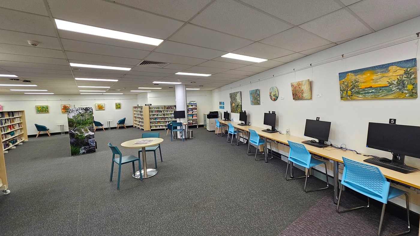 A library interior with a row of computer stations on the right, each with a monitor and a blue chair. The left side features bookshelves filled with books and a few teal armchairs. In the centre, there are small round tables with chairs. The walls display various paintings, and the ceiling has fluorescent lighting. The floor is carpeted in grey. A library interior with a row of computer stations on the right, each with a monitor and a blue chair. The left side features bookshelves filled with books and a few teal armchairs. In the centre, there are small round tables with chairs. The walls display various paintings, and the ceiling has fluorescent lighting. The floor is carpeted in grey.