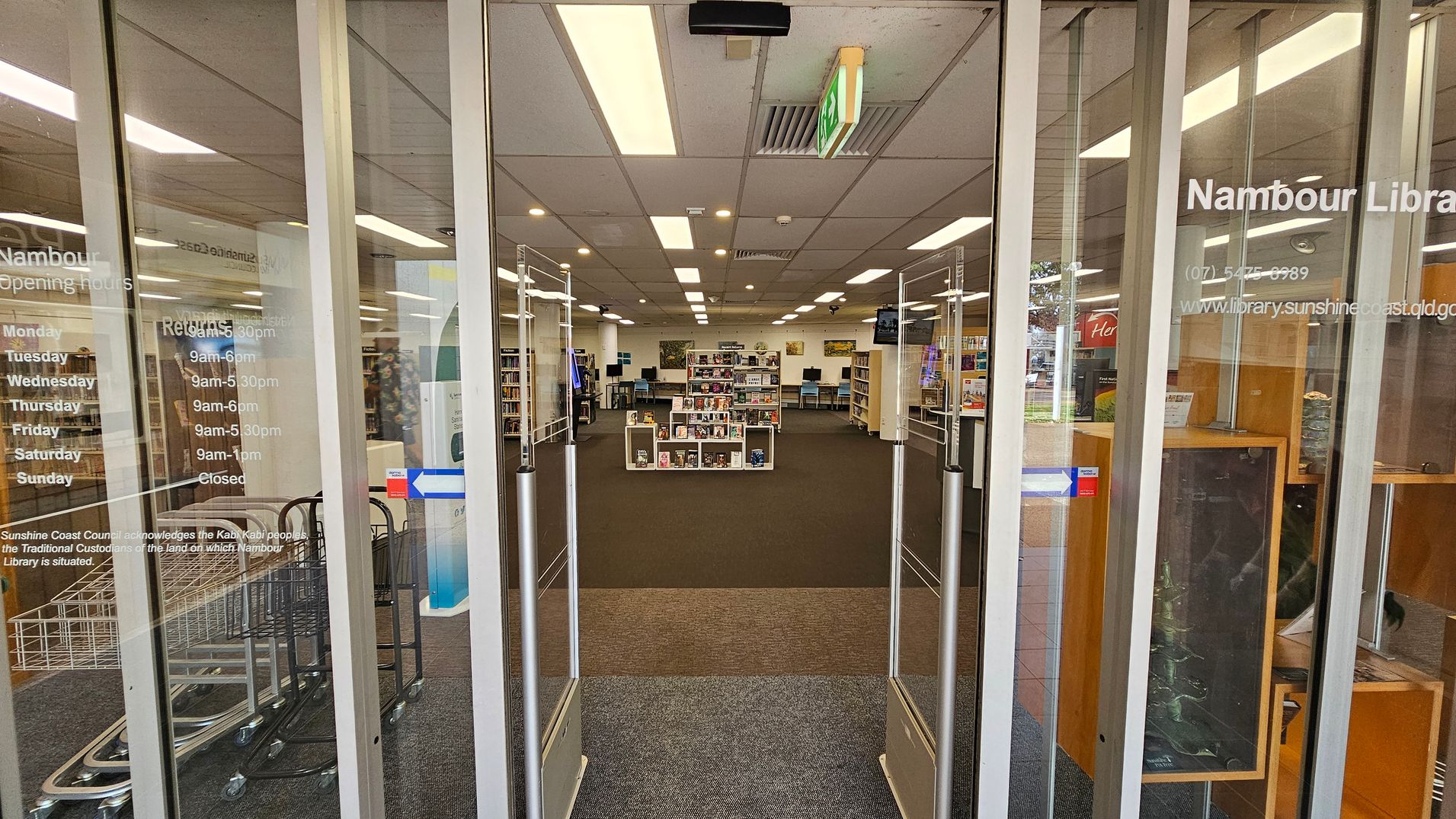 Glass entrance doors to a library with opening hours listed on the left. Inside, the library has a carpeted floor and a grid of fluorescent lights on the ceiling. In the centre, there are bookshelves filled with books and media. To the right, there is a wooden display case. In the background, more shelves and computer stations are visible. An exit sign hangs from the ceiling. Glass entrance doors to a library with opening hours listed on the left. Inside, the library has a carpeted floor and a grid of fluorescent lights on the ceiling. In the centre, there are bookshelves filled with books and media. To the right, there is a wooden display case. In the background, more shelves and computer stations are visible. An exit sign hangs from the ceiling.