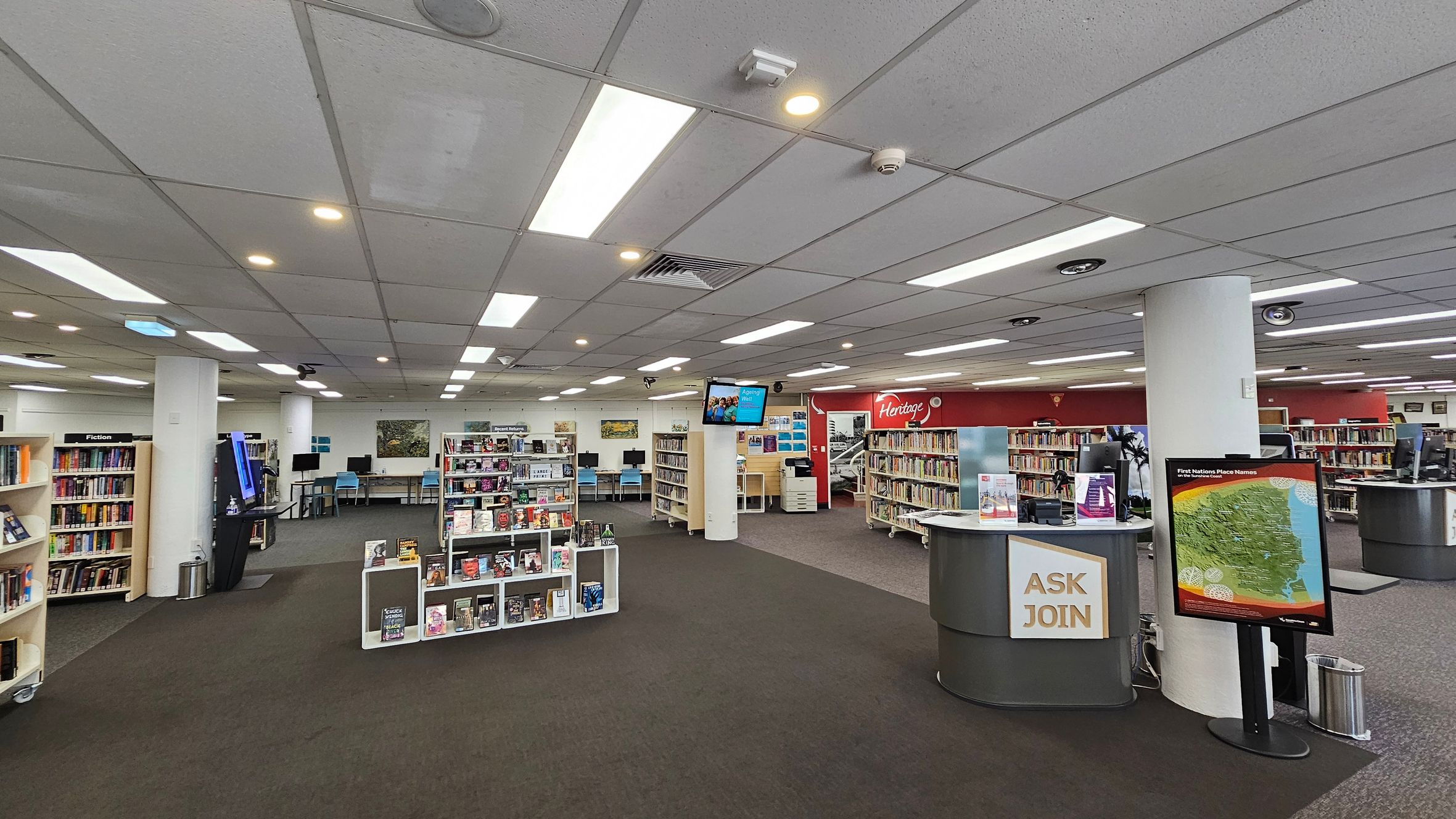 A spacious library interior with a grey-tiled ceiling and bright fluorescent lights. In the foreground, a circular desk with a sign reading "ASK JOIN" and a map display. To the left, bookshelves labelled "Fiction" and a digital kiosk. In the centre, more bookshelves and a TV screen mounted on a pillar. The background features computer stations with blue chairs and a red wall with "Heritage" signage. The floor is carpeted in dark green and grey. A spacious library interior with a grey-tiled ceiling and bright fluorescent lights. In the foreground, a circular desk with a sign reading "ASK JOIN" and a map display. To the left, bookshelves labelled "Fiction" and a digital kiosk. In the centre, more bookshelves and a TV screen mounted on a pillar. The background features computer stations with blue chairs and a red wall with "Heritage" signage. The floor is carpeted in dark green and grey.
