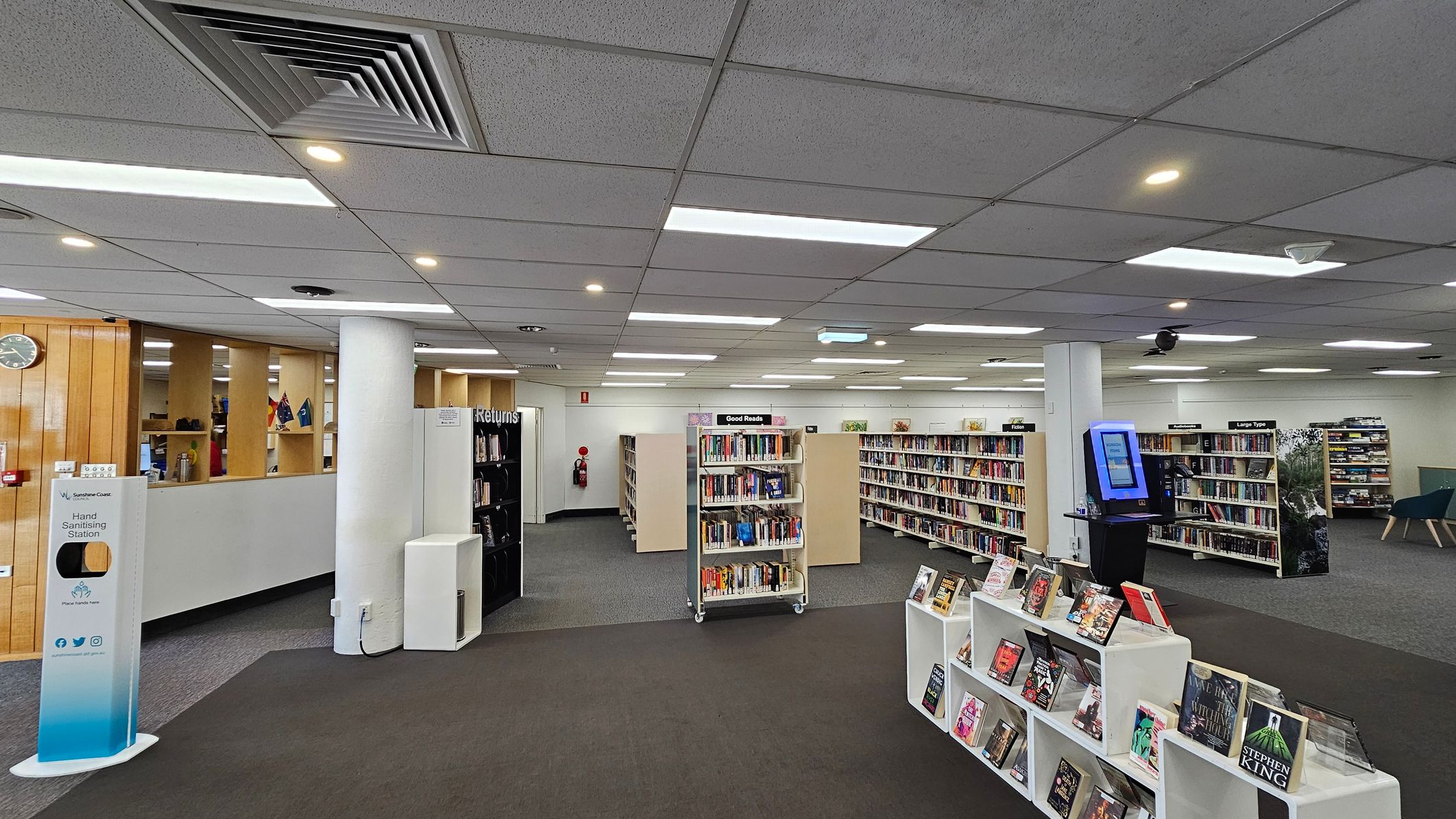 A library interior with a grey carpeted floor and a ceiling with recessed lighting and air vents. In the foreground, there are white shelves displaying books. In the middle, several bookcases are filled with books, labelled "Good Reads" and "Large Type". On the right, a digital kiosk stands near a column. On the left, a hand sanitising station is positioned near a wooden partition with a clock and small flags. A "Returns" section is visible in the background. A library interior with a grey carpeted floor and a ceiling with recessed lighting and air vents. In the foreground, there are white shelves displaying books. In the middle, several bookcases are filled with books, labelled "Good Reads" and "Large Type". On the right, a digital kiosk stands near a column. On the left, a hand sanitising station is positioned near a wooden partition with a clock and small flags. A "Returns" section is visible in the background.