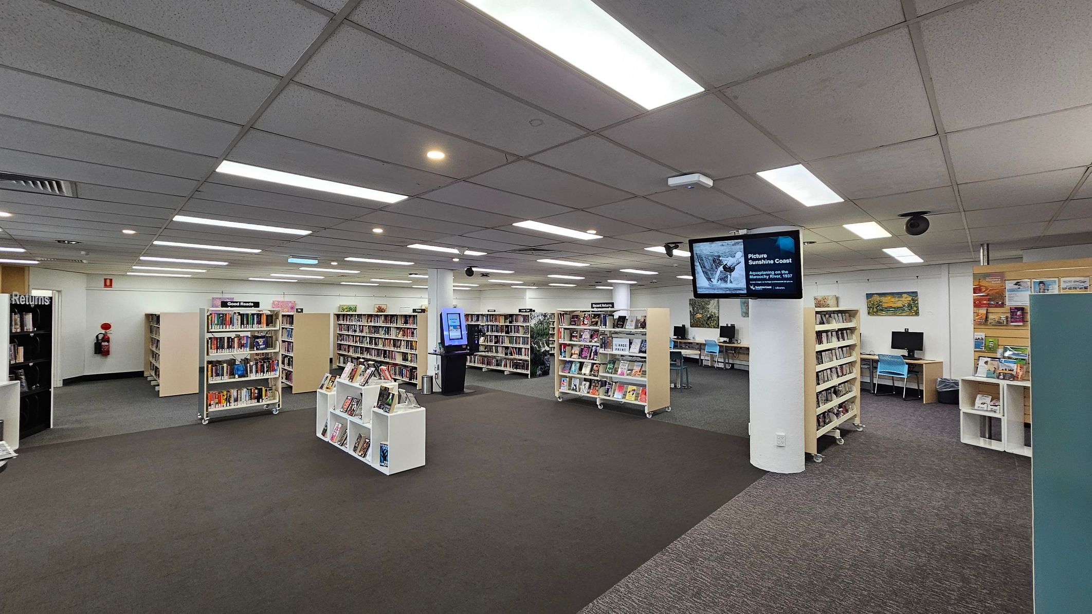 A library interior with grey carpeted flooring and a grid-patterned ceiling with fluorescent lights. In the centre, there are several bookshelves filled with books. On the right, a digital screen is mounted on a pillar, and computer stations with blue chairs are visible. On the left, a book return area is marked. The walls have posters and artwork, and a fire extinguisher is mounted near the corner. A library interior with grey carpeted flooring and a grid-patterned ceiling with fluorescent lights. In the centre, there are several bookshelves filled with books. On the right, a digital screen is mounted on a pillar, and computer stations with blue chairs are visible. On the left, a book return area is marked. The walls have posters and artwork, and a fire extinguisher is mounted near the corner.